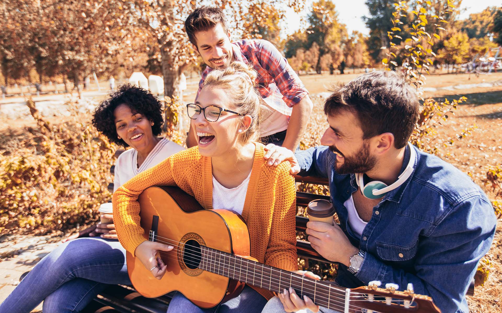 About Our Agency - Portrait of a Cheerful Group of Friends Sitting on a Bench in the Park During the Fall on a Sunny Day While One Friend Plays the Guitar